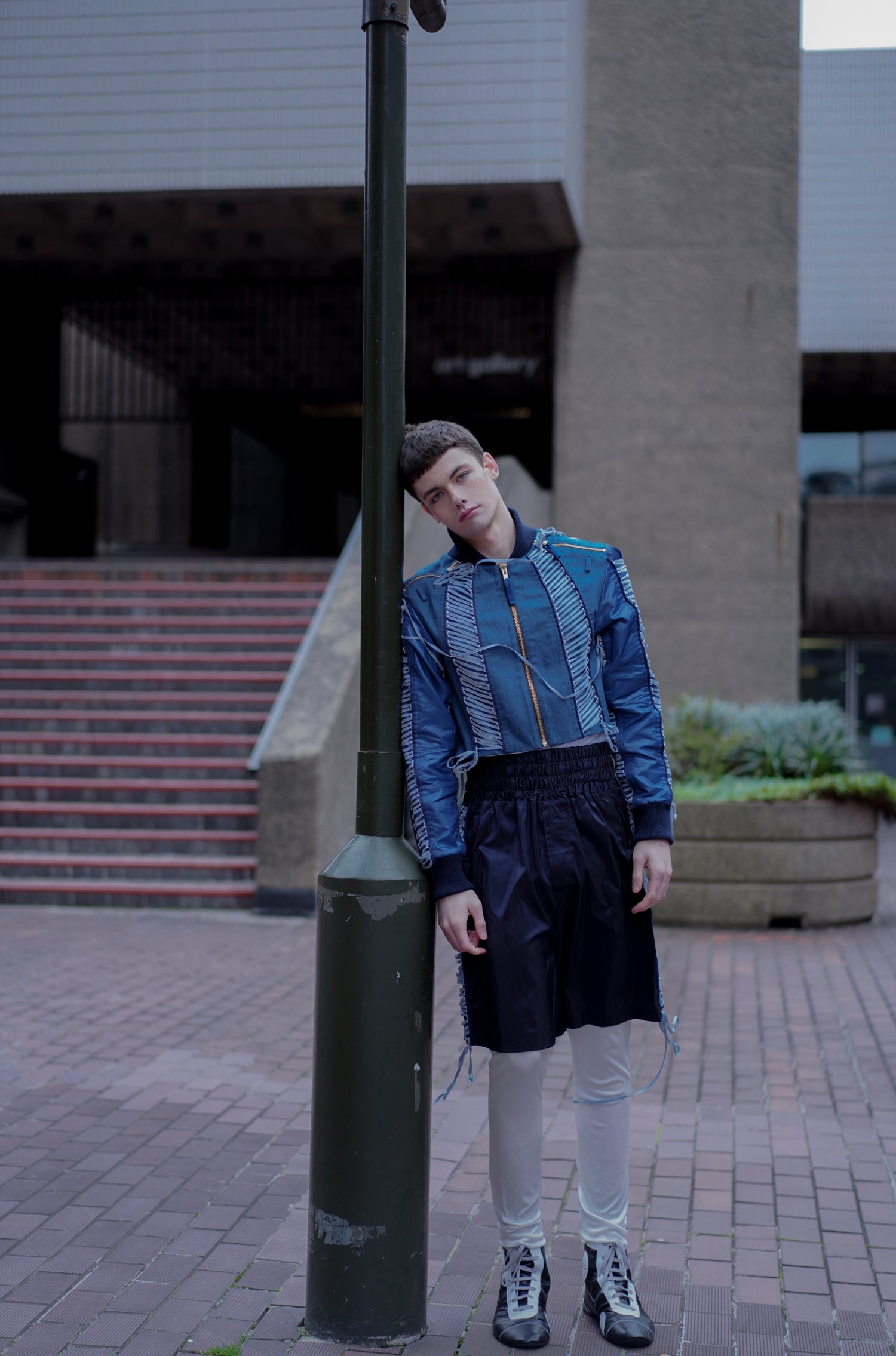Frontal shot of model wearing YYAtomic blue denim lacing jacket and navy blue boxing lacing shorts while leaning on a street lantern.