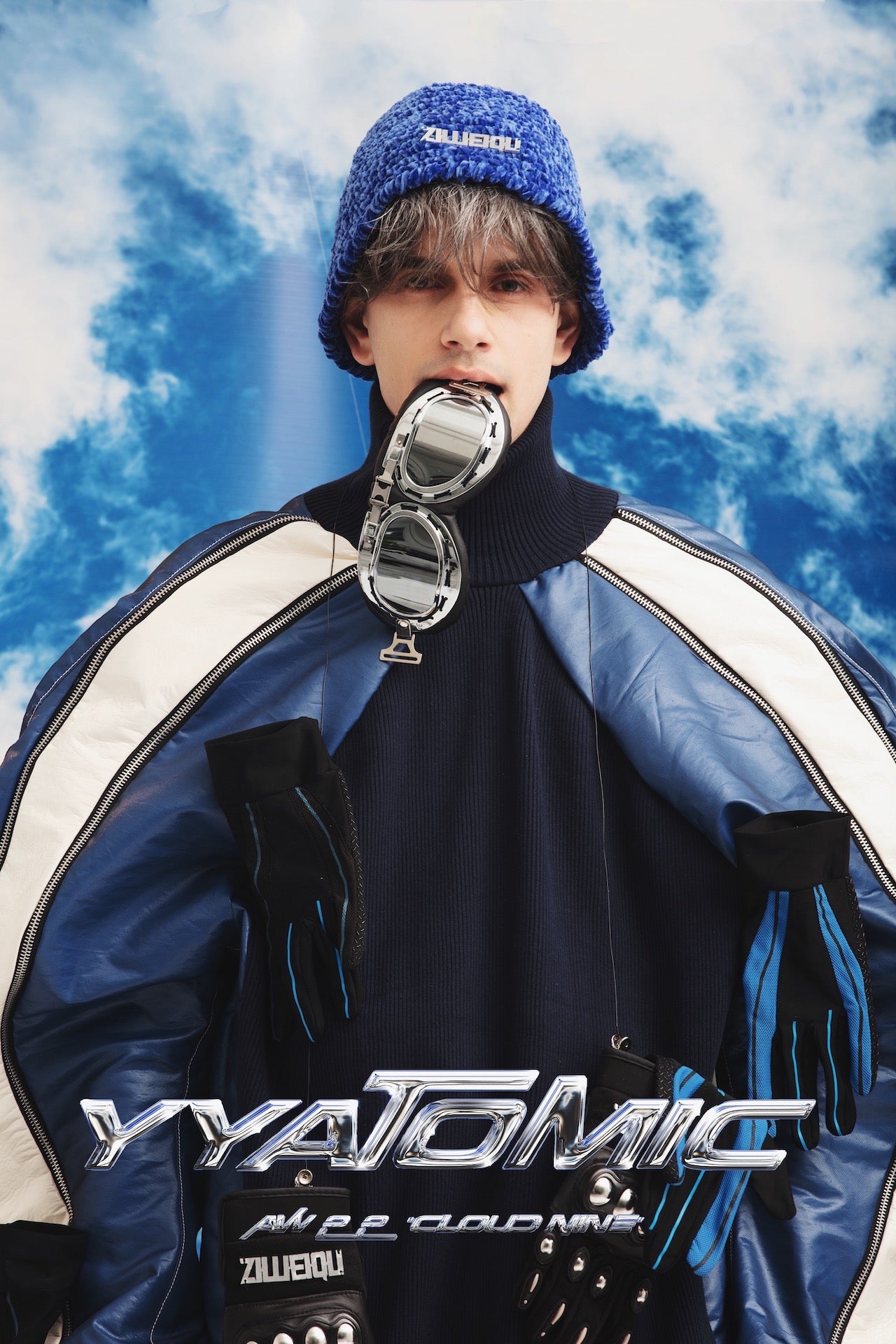 Frontal shot of model wearing YYAtomic's Raglan Sleeve Jumper, Blue Wool Bucket Hat and holding aviator goggles in his mouth.